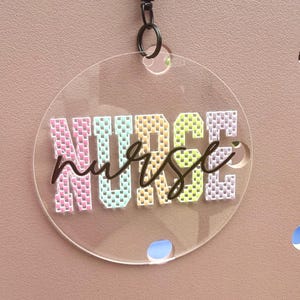 May include: A circular clear acrylic tag featuring the word NURSE in a blocky, multi-colored pixelated font with pink, blue, yellow, green, and purple letters. Overlaid in the center is the word nurse in a dark brown cursive script.