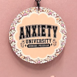 Może przedstawiać: Okrągła zawieszka do torby z falistym brzegiem, motywem kwiatowym i brzoskwiniowym środkiem. Napis ANXIETY UNIVERSITY HONORS PROGRAM wykonano czarnymi literami blokowymi. Zawieszka jest przymocowana do brudnoróżowej torby czarnym metalowym zapięciem.