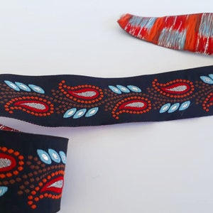 May include: A black ribbon with a paisley pattern in red, silver, and light blue. The ribbon has a repeating design of paisley shapes and small dots. The end of the ribbon has a red, orange, and silver striped pattern.