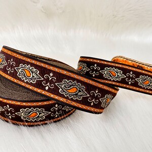 May include: Decorative ribbon with a dark brown base, featuring an embroidered paisley design in orange and gold. The ribbon is rolled and displayed on a white, fluffy surface, showcasing its intricate details.