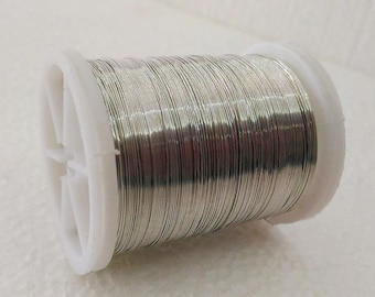 Può includere: Una bobina di filo color argento, avvolta strettamente attorno a un'anima di plastica bianca. Il filo appare liscio e riflettente, suggerendo una composizione metallica. La bobina è posizionata ad angolo, mostrando le spire ben ordinate.