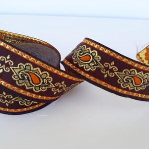 May include: A roll of decorative ribbon featuring a repeating paisley design. The ribbon is primarily brown with gold and orange accents. The edges are trimmed with gold, and the paisley shapes are outlined in gold.