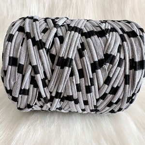 May include: A cylindrical ball of gray and black striped t-shirt yarn. The yarn is made of thick, soft fabric strips, ideal for crafting projects like crochet or knitting. The yarn is displayed on a white, fluffy surface.