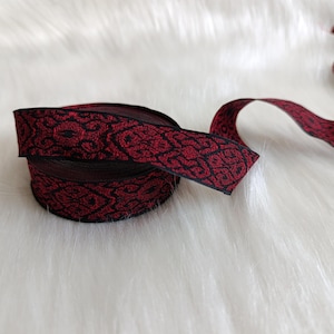Peut inclure: Un rouleau de ruban décoratif avec un motif noir et rouge répété. Le ruban est déroulé, avec une partie enroulée et le reste s'étendant. Le ruban est posé sur une surface blanche et duveteuse.