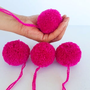 Könnte beinhalten: Vier flauschige, leuchtend rosa Pompons mit langen Schnüren. Die Pompons sind aus Garn und haben einen Durchmesser von etwa 7,5 cm.