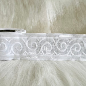 May include: A roll of white decorative ribbon with an embroidered scroll design. The ribbon is made of a soft, textured fabric and is ideal for crafts, sewing, or adding a decorative touch to gifts. The ribbon is approximately 2 inches wide.