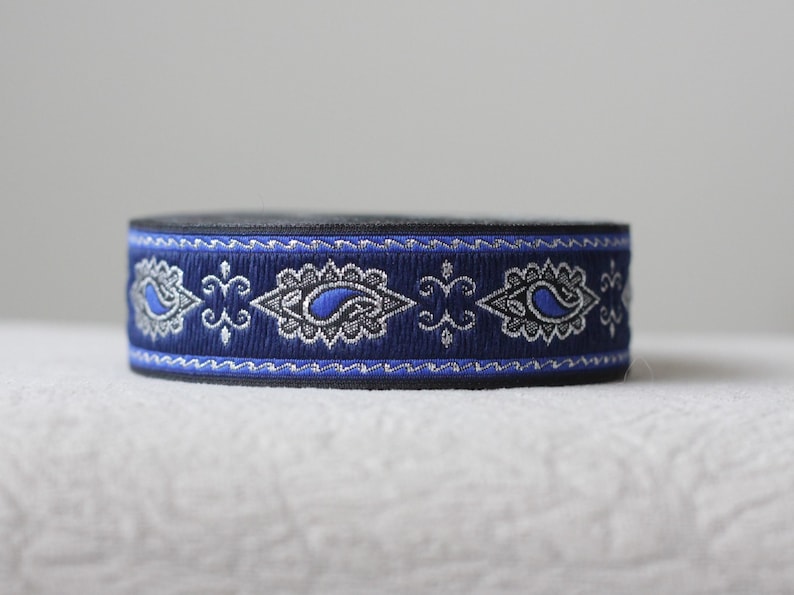 Puede incluir: Cinta de adorno azul y plata con estampado paisley y respaldo negro. La cinta tiene un patr&oacute;n repetitivo de formas paisley y un borde ondulado.