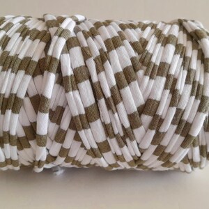 May include: A cylindrical roll of white and olive green striped fabric, likely t-shirt yarn. The yarn is tightly wound, creating a textured surface. This material is suitable for various crafting projects, such as crochet or knitting.