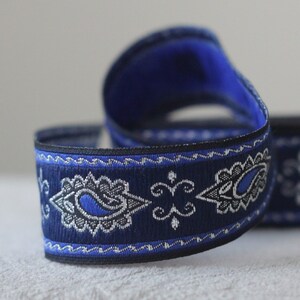 Puede incluir: Cinta de adorno azul y plata con estampado paisley y borde negro. La cinta tiene un patr&oacute;n repetido de formas paisley y una l&iacute;nea ondulada.