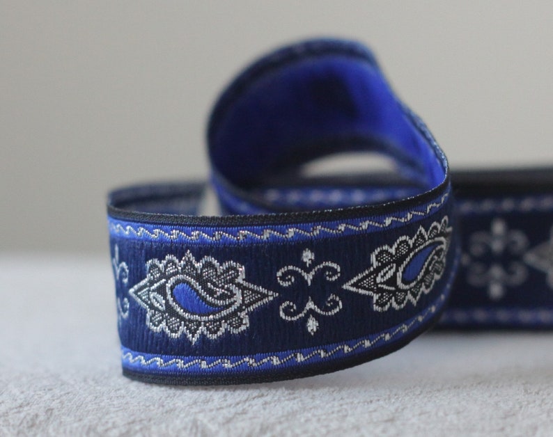 Puede incluir: Cinta de adorno azul y plata con estampado paisley y borde negro. La cinta tiene un patr&oacute;n repetido de formas paisley y una l&iacute;nea ondulada.