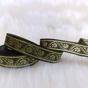 Op de afbeelding: Een rol olijfgroen geweven lint met een herhalend crèmekleurig bloemenpatroon met krullende rankdetails. De stoffen bies heeft een getextureerd oppervlak en afgewerkte randen, met een doorlopend decoratief ontwerp over de gehele lengte.