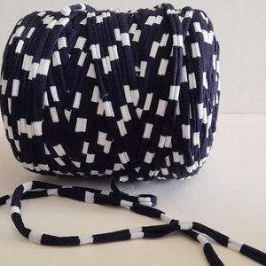 May include: A large ball of navy blue and white fabric with a repeating pattern of white rectangles. A length of the same fabric extends from the ball, forming a striped cord. The fabric appears to be a knitted or woven material, suitable for crafting.