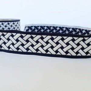 May include: A roll of decorative ribbon with a woven basket weave pattern. The ribbon features a black border with a white and black design. The ribbon is likely used for crafting, sewing, or gift wrapping.