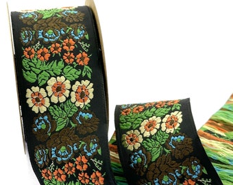 Könnte beinhalten: Eine Rolle schwarzes Stoffband mit einem gewebten Blumenmuster in Orange, Hellblau, Creme und Grün. Das Band zeigt ein sich wiederholendes Design aus Blüten und Blättern, mit einer ausgefransten Kante, die lose Fäden in passenden Farben zeigt.