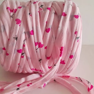 May include: A spool of pink fabric yarn with a repeating pattern of pink hearts and black floral accents. The yarn is tightly wound, with a loose end extending from the bottom. Ideal for crafting projects.