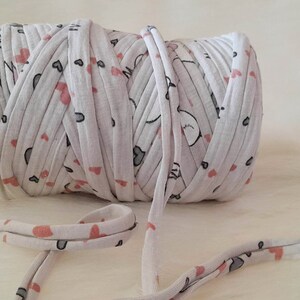 Puede incluir: Un rollo de hilo de tela blanco con un patrón repetido de corazones rosas y formas grises. Un hilo del hilo se extiende desde el rollo. Este hilo se utiliza probablemente para proyectos de artesanía y textiles. El hilo tiene un estampado de corazones.
