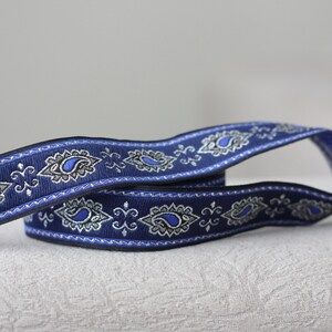 Puede incluir: Cinta de adorno azul y plata con estampado paisley y borde decorativo. La cinta est&aacute; doblada por la mitad y los extremos se superponen.
