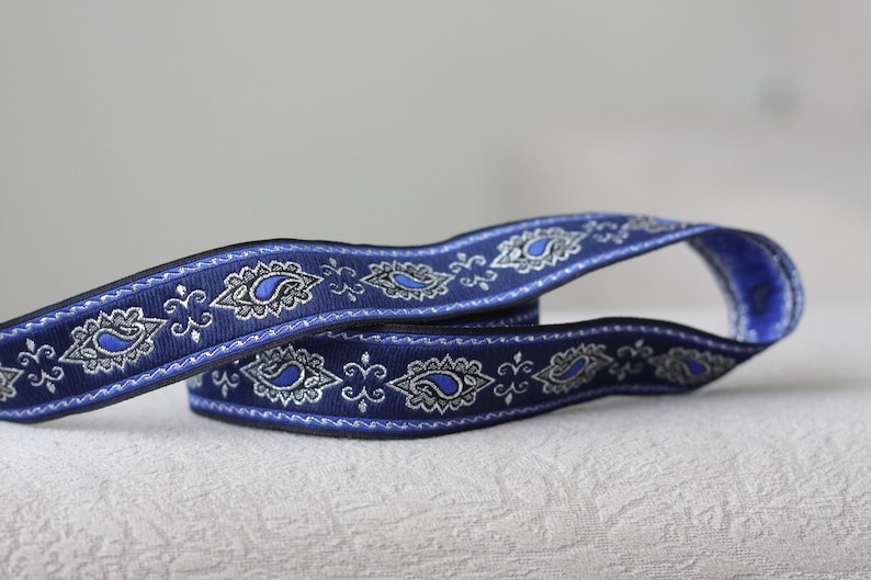 Puede incluir: Cinta de adorno azul y plata con estampado paisley y borde decorativo. La cinta est&aacute; doblada por la mitad y los extremos se superponen.