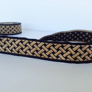 May include: Black and gold woven trim with a geometric pattern. The trim is approximately 1 inch wide.
