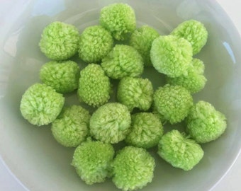 Può includere: Una ciotola bianca piena di pompon verde lime. I pompon sono rotondi e soffici, con una consistenza visibile. La ciotola curva e la luce creano un'immagine morbida e luminosa.