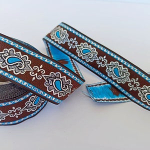 May include: Brown and blue decorative ribbon with a paisley design. The ribbon features a repeating pattern of blue paisley shapes, white and gray floral accents, and a blue zigzag border. A roll of the ribbon is visible.