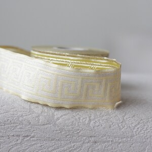 May include: A roll of white and gold trim with a geometric pattern. The trim is on a white textured surface.