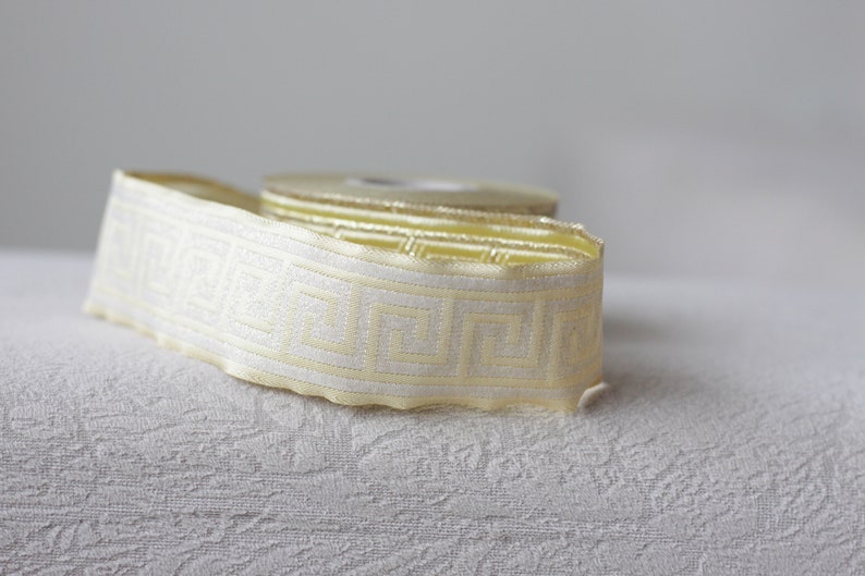 May include: A roll of white and gold trim with a geometric pattern. The trim is on a white textured surface.