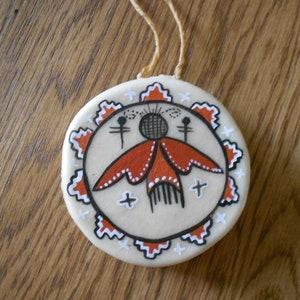 May include: A round, white, leather ornament with a painted design of a stylized figure with red wings and a black head. The ornament has a white cord for hanging.