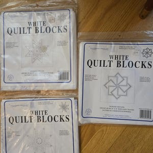 May include: Several packages of white quilt blocks are displayed, each sealed in clear plastic. The packages feature the text "WHITE QUILT BLOCKS" in bold letters, along with various star and geometric designs. The packages are for embroidery.
