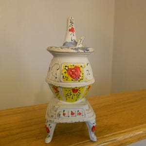 May include: A vintage, white cast iron stove with a decorative floral design. The stove features a yellow and red floral pattern on the body and legs. The stove has a tall chimney and is painted with red accents.