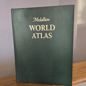 May include: A dark green hardcover book titled "Medallion WORLD ATLAS" in gold lettering. The book is standing upright on a wooden surface, with a neutral background. The book's cover is slightly worn.