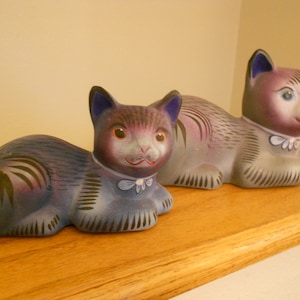 May include: Two ceramic cat figurines, one gray and one purple, with painted details and blue accents. The cats are lying down with their heads turned to the side.