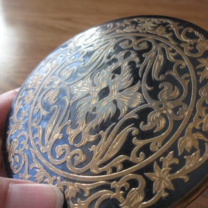 May include: A round, black compact with an ornate gold floral design. The compact's lid features a detailed pattern of swirling leaves and flowers, with a central emblem. The design is set against a black background.