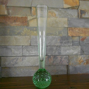 May include: A tall, clear glass vase with a green, bubbled base. The vase is narrow and cylindrical, with a rounded bottom. The base is covered in small, evenly spaced bubbles.
