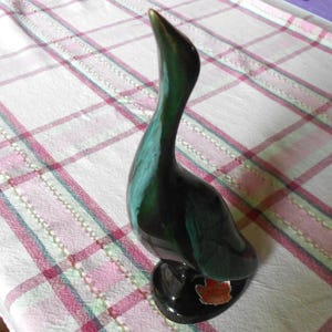 May include: A ceramic duck figurine with a glossy, dark green and teal finish. The duck has a long, curved neck and a small, round base. The figurine has a small, orange and brown accent on the base.
