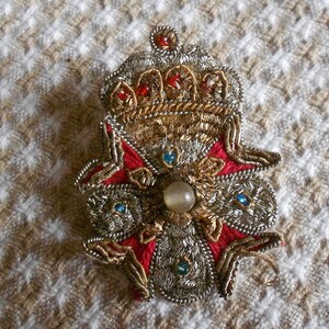 May include: A gold and silver embroidered brooch with a red and white flower design and a pearl-like center. The brooch features a crown at the top.