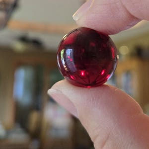 May include: A small, spherical, translucent dark red glass marble. The surface of the marble is smooth and polished, reflecting light with bright highlights. The deep crimson color is consistent throughout the orb, showing internal clarity.