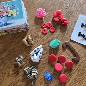 May include: A rectangular metal tin with a floral print, alongside a collection of assorted sewing buttons in various shapes, sizes, and colors including red, blue, white, green, and brown, plus two carded sets of buttons labeled le chic and Boutique.