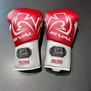 Puede incluir: Par de guantes de boxeo Rival RS100 rojos y blancos. Los guantes tienen una parte superior roja con un logotipo blanco y la palabra "RIVAL". La zona de la muñeca es plateada con el texto "RS100 SPARRING GLOVE".