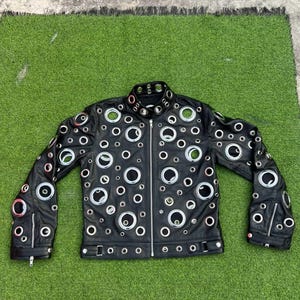 May include: Black leather jacket with numerous silver grommets of varying sizes. The jacket features a zip-up front, two zippered pockets, and a classic collar. The sleeves have zippers at the cuffs. The jacket is displayed on a green surface.