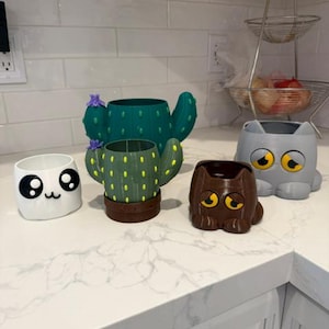 May include: A collection of five small planters including a white panda-faced pot, a dark green cactus-shaped pot, a light green cactus-shaped pot with yellow dots, a brown cat-shaped pot, and a gray cat-shaped pot with large yellow eyes.