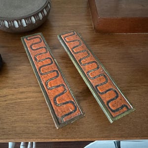 May include: Two vintage brass door handles with a geometric orange and brown design. The handles are rectangular and have a smooth, polished finish.