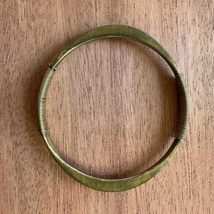 May include: A brass-toned bangle bracelet with a simple, elegant design. The bracelet features a smooth, curved band with a coiled wire detail on each side. The bracelet is approximately 2.5 inches in diameter.