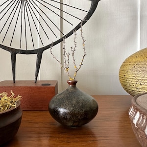 May include: A small, round, dark-toned ceramic vase with a smooth, glossy finish and a narrow neck. The vase contains a thin, bare, light-colored twig with small yellow lichen spots, standing upright in the opening of the vessel.