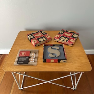 May include: Three boxes with red and black geometric patterns and the word "HAMMER" printed on them. A gray ceramic letter "S" is displayed on a wooden table with white metal legs. The letter is in a box with a red and black geometric pattern.