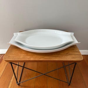 May include: Two stacked white ceramic serving platters with a smooth, glossy finish. The platters feature a distinct elongated, leaf-like shape with pointed ends and raised edges, creating a clean, uniform appearance for serving food items.