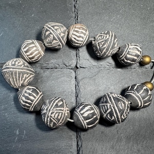 Könnte beinhalten: Eine Kette aus dunkelgrauen, kugelförmigen Perlen mit weißen, eingravierten geometrischen Mustern. Die Kette besteht aus zehn texturierten, tonartigen Perlen und zwei glatten, goldfarbenen Metallkugeln, die auf einer hellen Schnur aufgereiht sind.