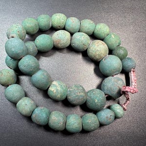May include: A necklace or bracelet featuring a strand of turquoise beads with a weathered look. The round beads are strung on a pinkish-red cord. The beads vary in size and shade, creating a unique, aged aesthetic.