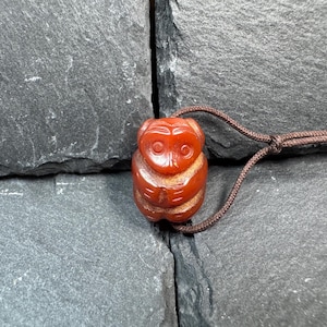 May include: A reddish-brown owl pendant with a brown cord. The owl is carved with detailed features, including eyes and feathers. The pendant is made of a smooth, polished material and is set against a dark, textured background.