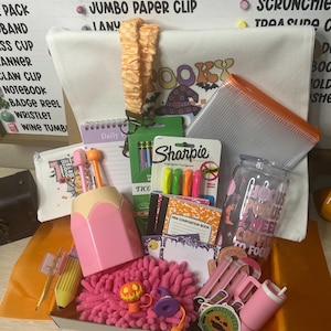 May include: A gift box containing a white tote bag with a spooky graphic, a pink pencil-shaped cup, a checkered orange wristlet, a clear zipper pouch, a glass tumbler with text, Sharpie highlighters, mini composition books, and various stationery items.